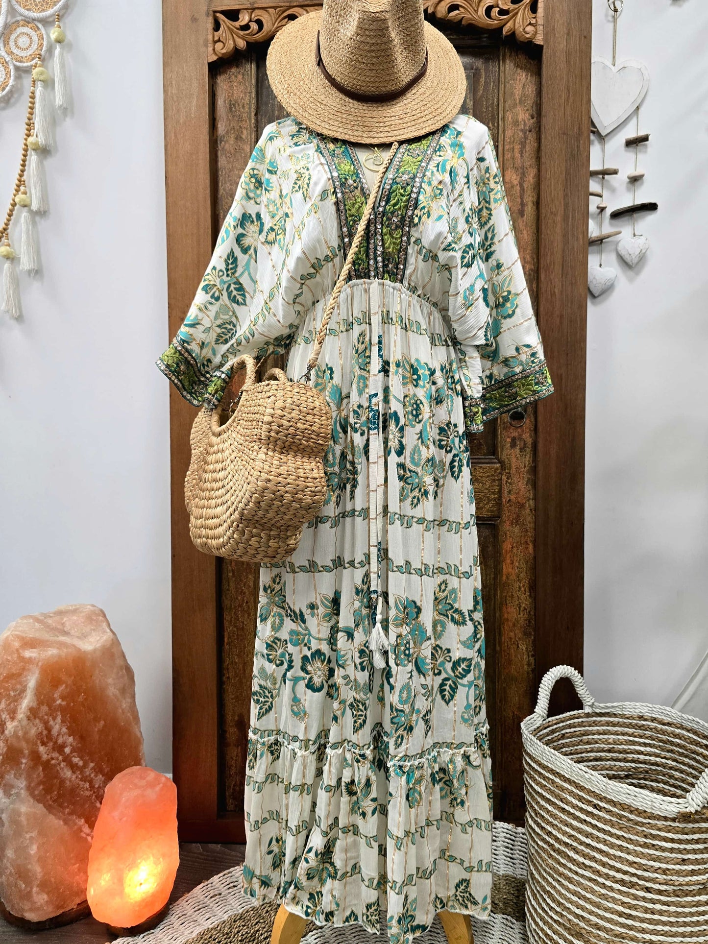 A bohemian-style tiered maxi dress with a green and teal floral block print on an off-white base, styled on a mannequin with a straw hat, woven shoulder bag, and a matching woven basket bag, set against a rustic wooden door with decorative wall hangings and salt lamps.