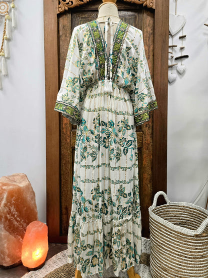 A bohemian-style tiered maxi dress with a green and teal floral print on an off-white base, displayed on a mannequin in front of a dark wooden door. The dress features a V-neckline with green embroidered detailing and wide three-quarter sleeves.