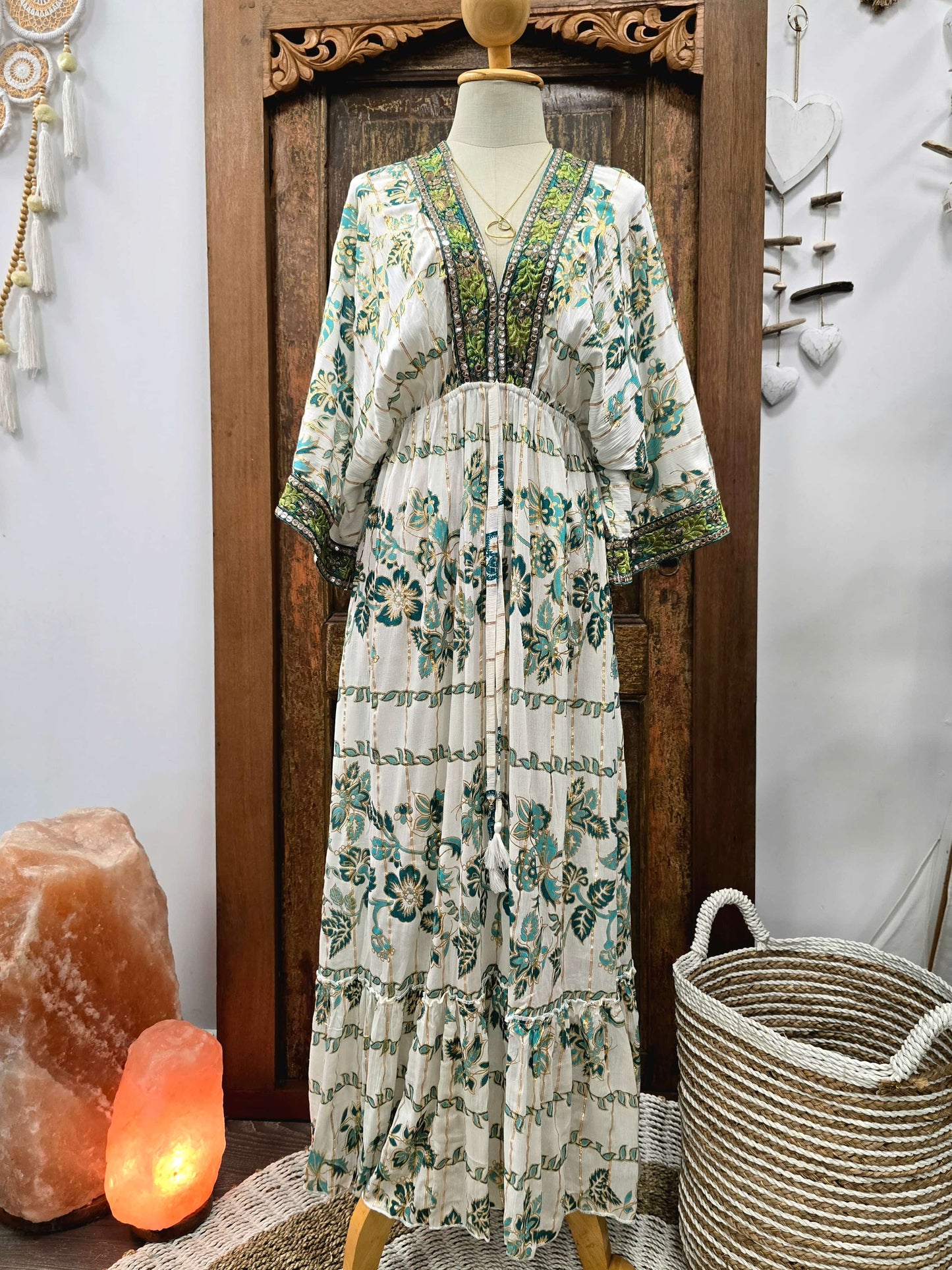 A bohemian-style maxi dress with a green and teal floral print on an off-white base, displayed on a mannequin in front of a carved wooden door. The dress features a V-neckline with green beaded trim and wide, elbow-length sleeves.