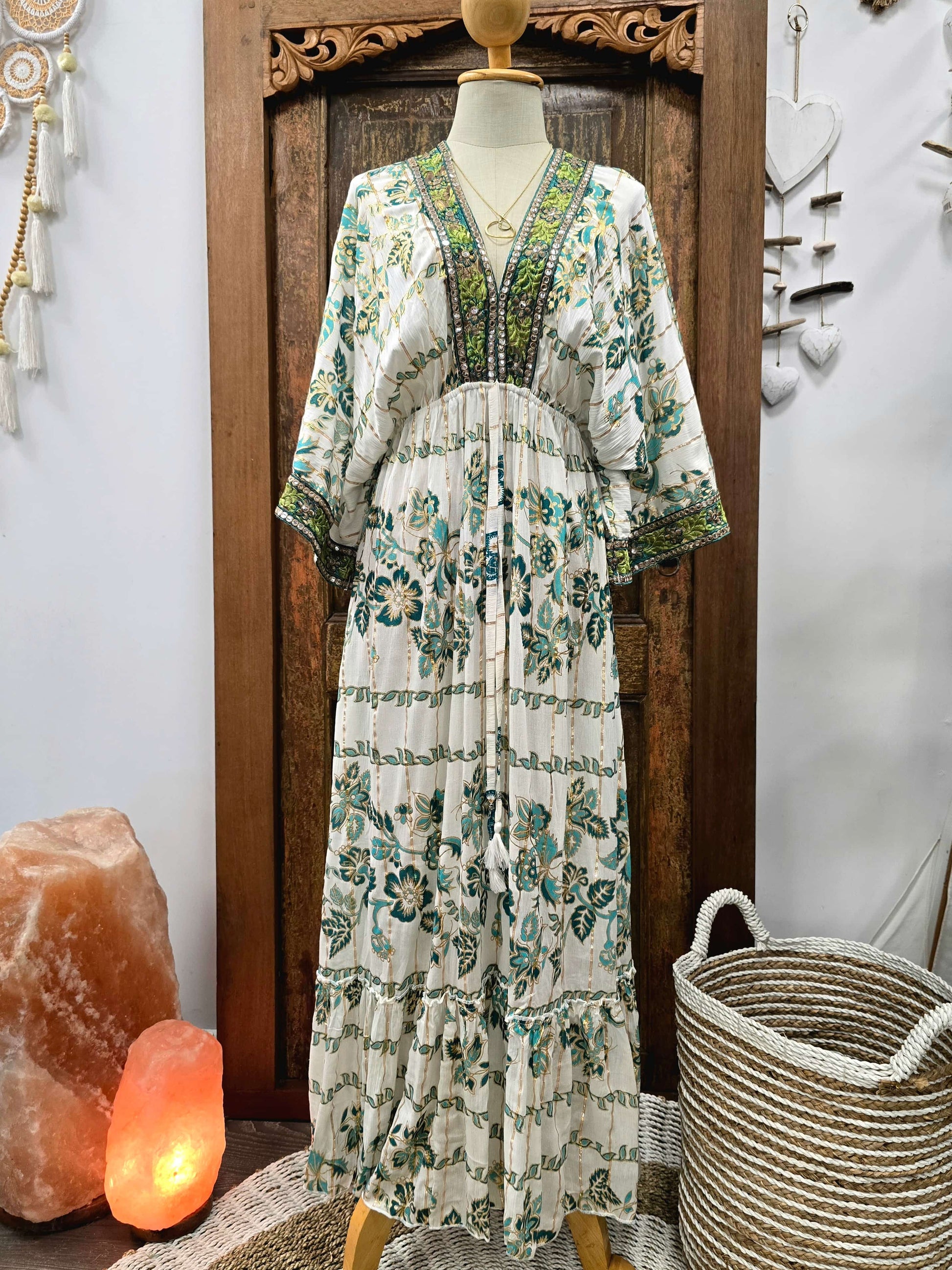 A bohemian-style maxi dress with a green and teal floral print on an off-white base, displayed on a mannequin in front of a carved wooden door. The dress features a V-neckline with green beaded trim and wide, elbow-length sleeves.