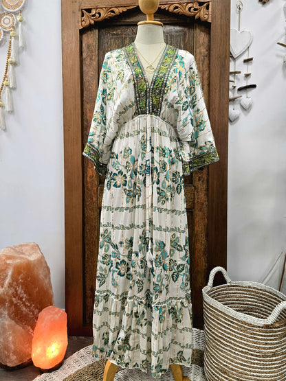 A bohemian-style maxi dress with a green and teal floral print on an off-white base, displayed on a mannequin in front of a carved wooden door. The dress features a V-neckline with green beaded trim and wide, elbow-length sleeves.