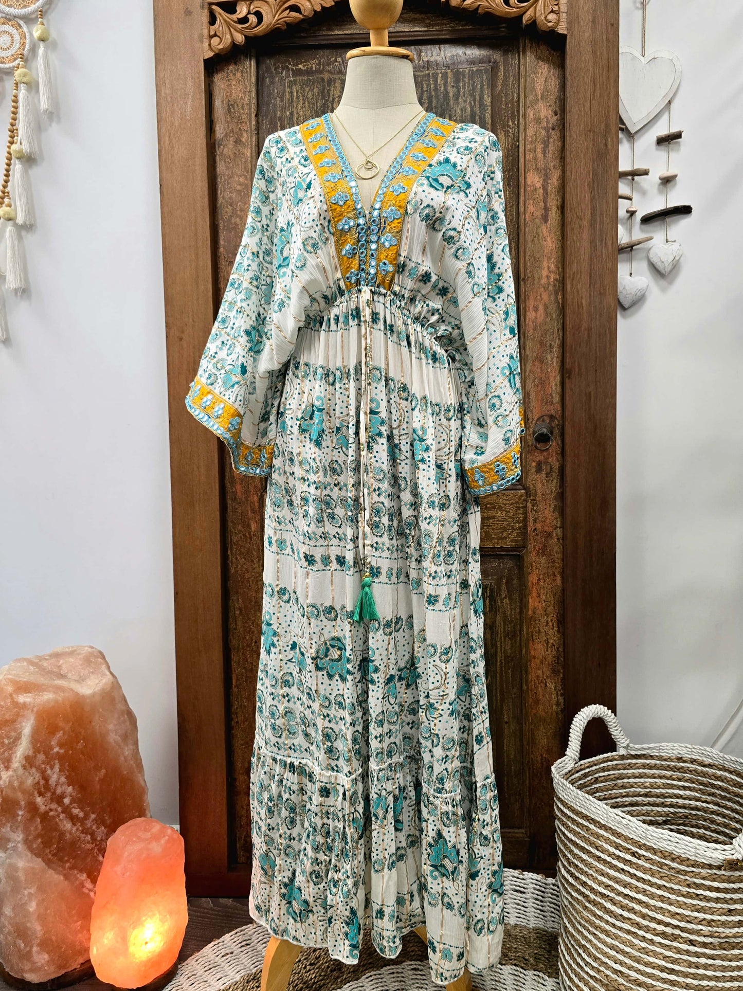 A bohemian-style maxi dress displayed on a mannequin torso, featuring a white base with teal and yellow floral patterns, set against a rustic wooden door background with decorative props.