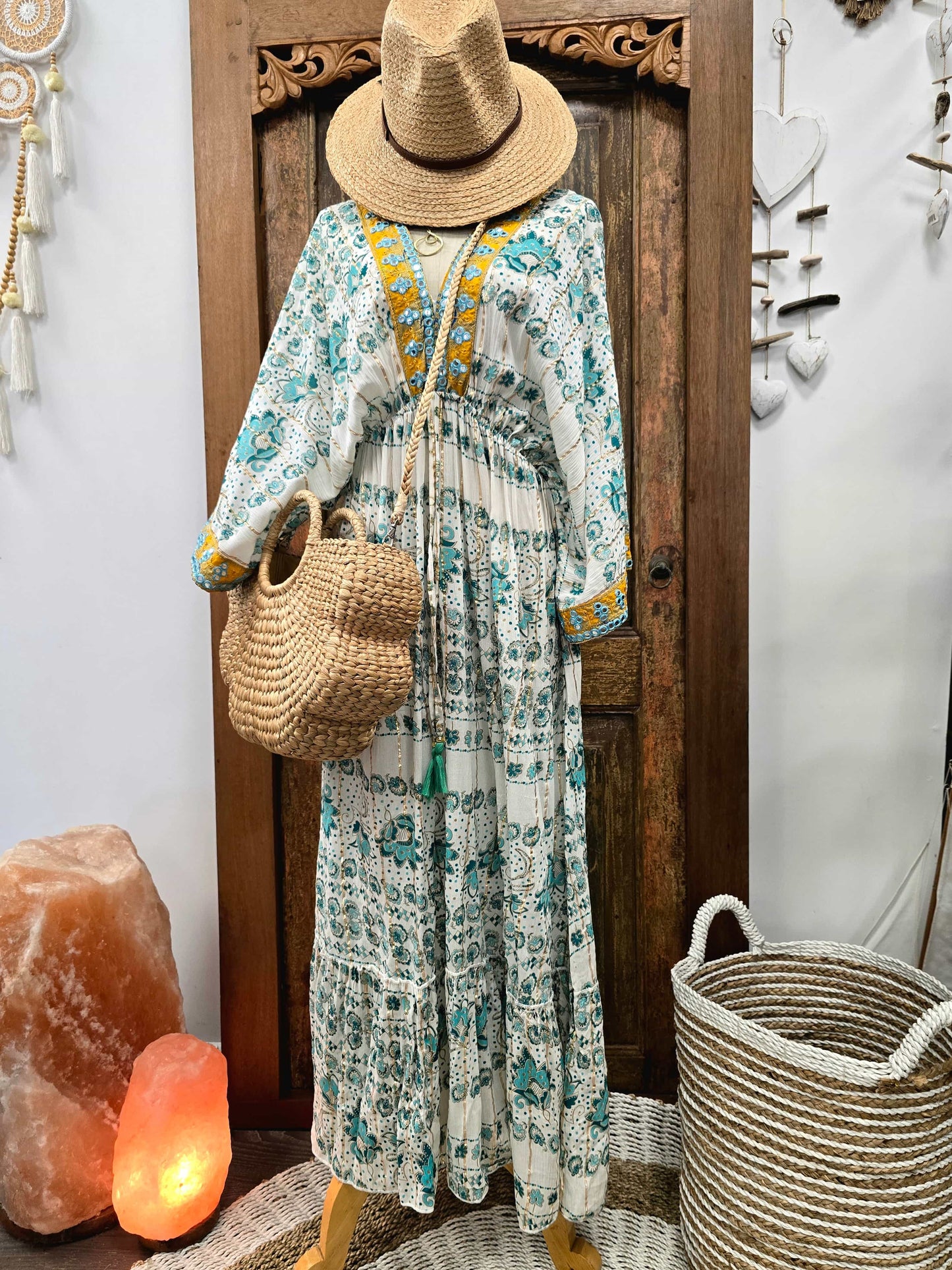 A bohemian-style long-sleeve maxi dress with a white base and teal floral/ornamental print, styled on a mannequin with a straw hat, woven tote bag, and yellow embroidered neckline details. The mannequin is posed in front of a carved wooden door with decorative home accessories like a salt lamp, woven basket, and wall hanging.