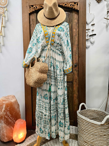 A bohemian-style long-sleeve maxi dress with a white base and teal floral/ornamental print, styled on a mannequin with a straw hat, woven tote bag, and yellow embroidered neckline details. The mannequin is posed in front of a carved wooden door with decorative home accessories like a salt lamp, woven basket, and wall hanging.
