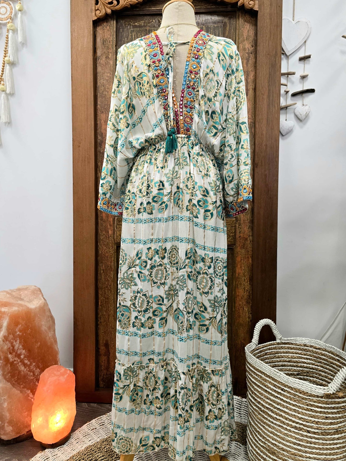The back of a long-sleeve bohemian maxi dress with a multicolor floral and geometric print on a cream base, displayed on a mannequin in front of a dark wooden door. The dress features a colorful embroidered V-neckline and a matching tie belt.