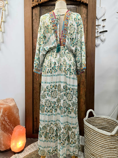 The back of a long-sleeve bohemian maxi dress with a multicolor floral and geometric print on a cream base, displayed on a mannequin in front of a dark wooden door. The dress features a colorful embroidered V-neckline and a matching tie belt.