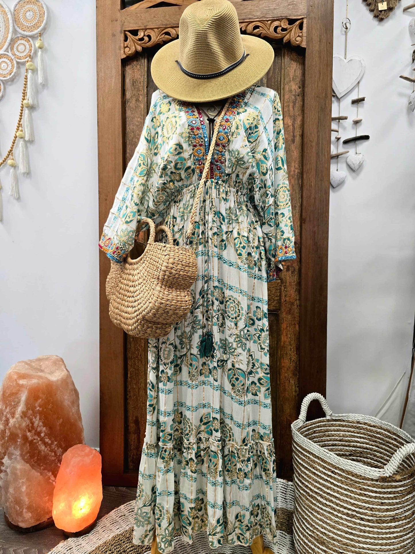 A bohemian-style long-sleeve maxi dress with a blue, green, and orange paisley pattern on a cream background, styled on a mannequin with a straw hat, woven shoulder bag, and standing in front of a carved wooden door.