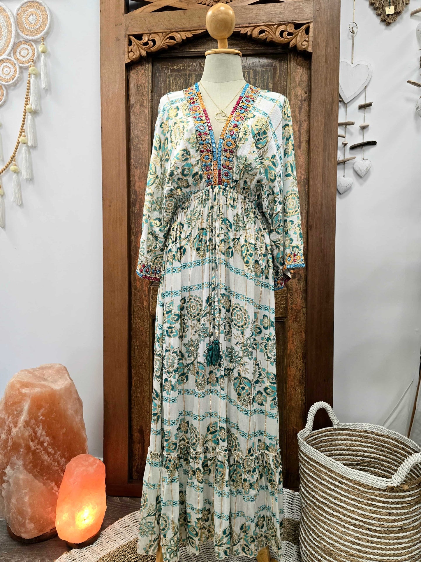 A long bohemian maxi dress with a white base and teal, green, and orange floral patterns, displayed on a mannequin in front of a carved wooden door.