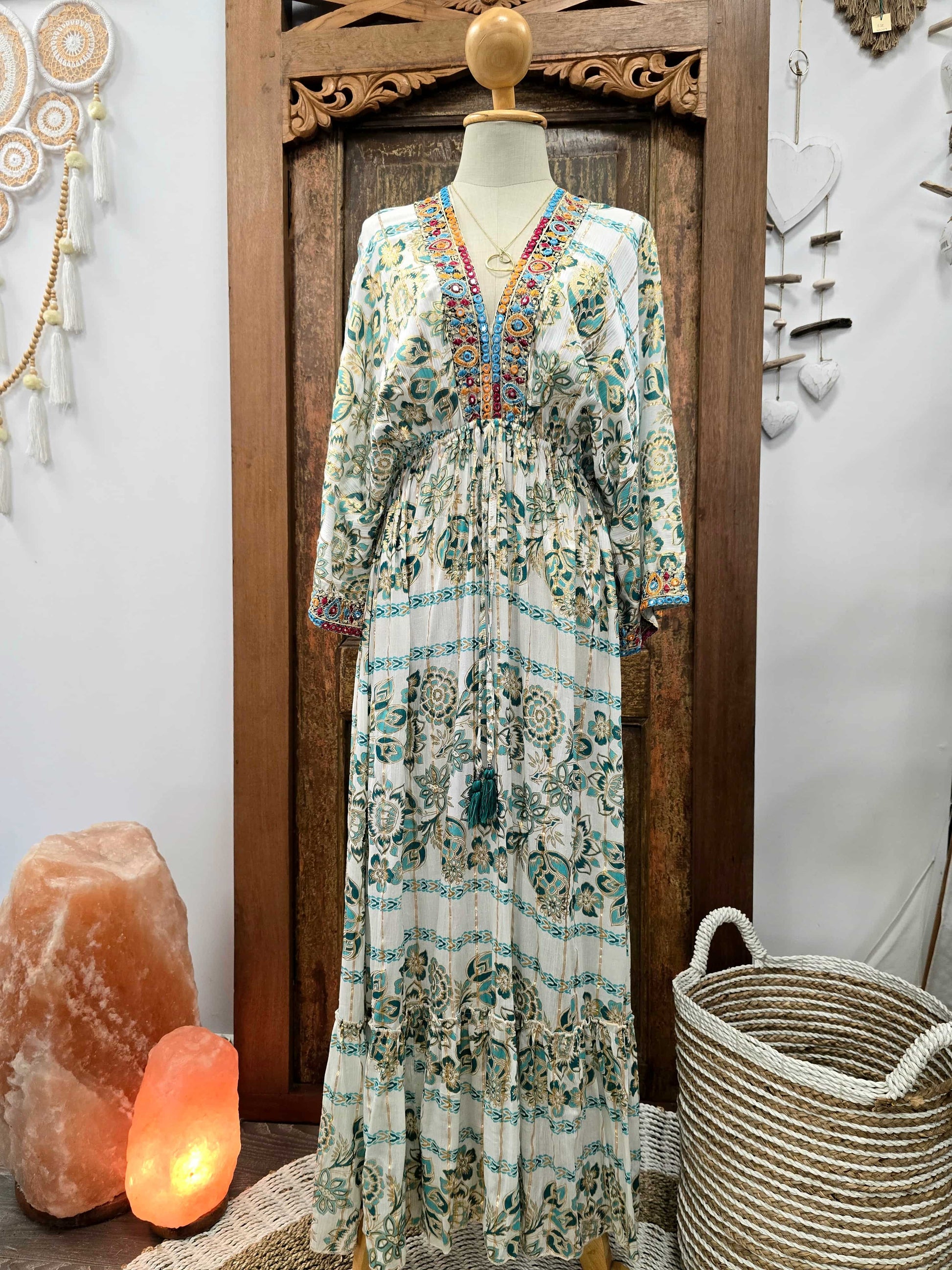 A long bohemian maxi dress with a white base and teal, green, and orange floral patterns, displayed on a mannequin in front of a carved wooden door.