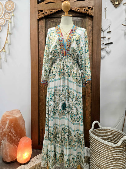 A long bohemian maxi dress with a white base and teal, green, and orange floral patterns, displayed on a mannequin in front of a carved wooden door.