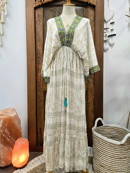 A bohemian-style maxi kaftan dress displayed on a mannequin in front of a wooden door. The dress is cream-colored with green and gold embroidered patterns, featuring a drawstring waist and tiered skirt.