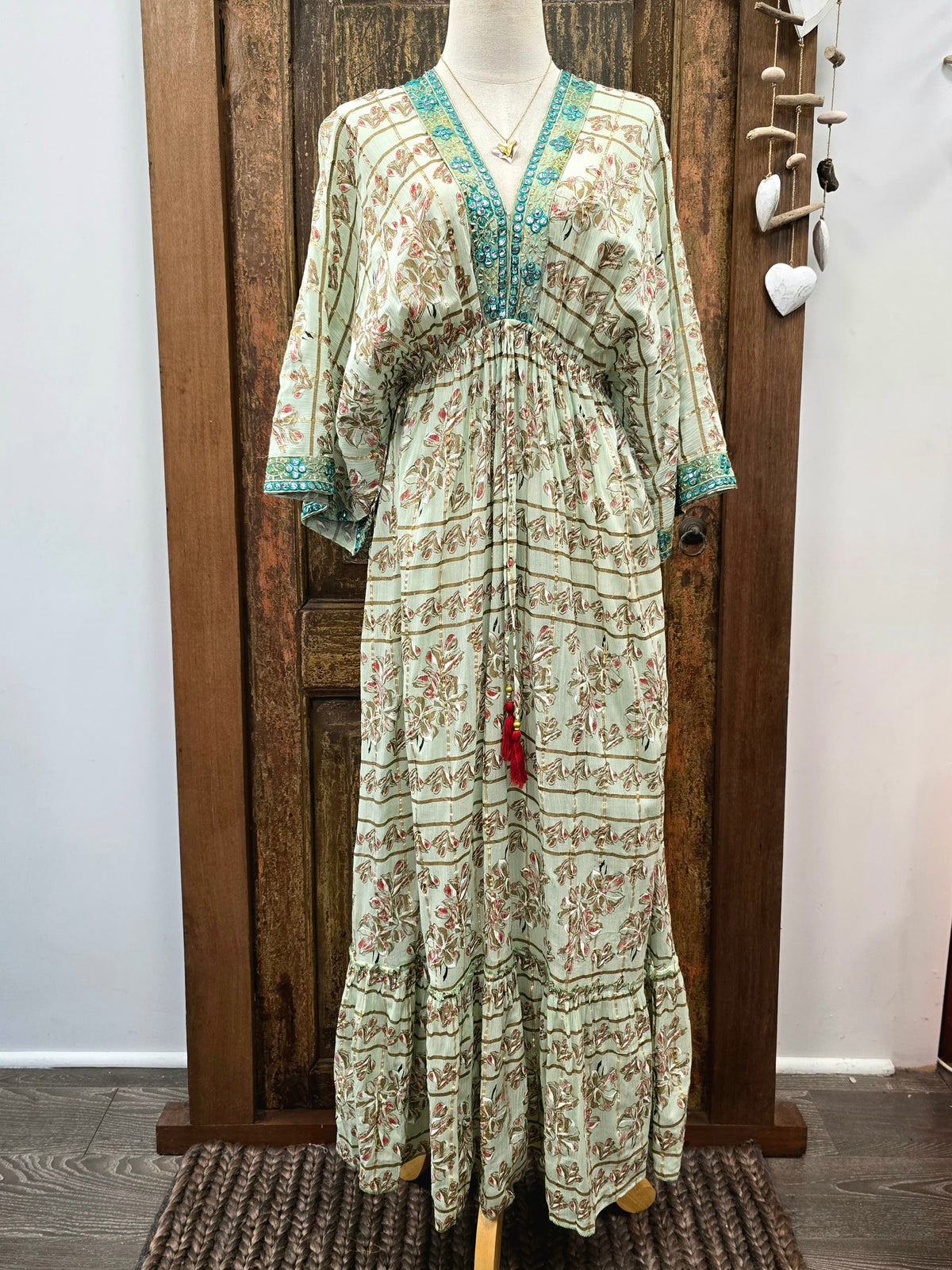 A bohemian-style maxi dress displayed on a mannequin in front of a rustic wooden door. The dress has a light sage green base with a brown floral pattern, turquoise blue embroidered trim along the V-neckline and cuffs, and a small red tassel hanging at the waist.
