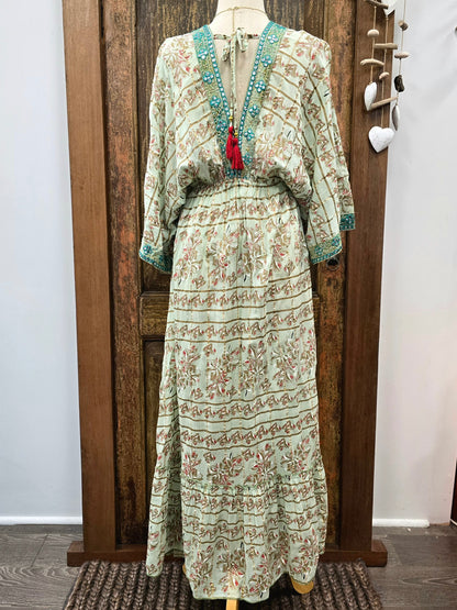 A bohemian-style maxi dress displayed on a mannequin in front of a rustic wooden door. The dress has a light sage green base with a brown floral pattern and turquoise blue embroidered trim along the V-neckline and sleeves, finished with a red tassel accent.