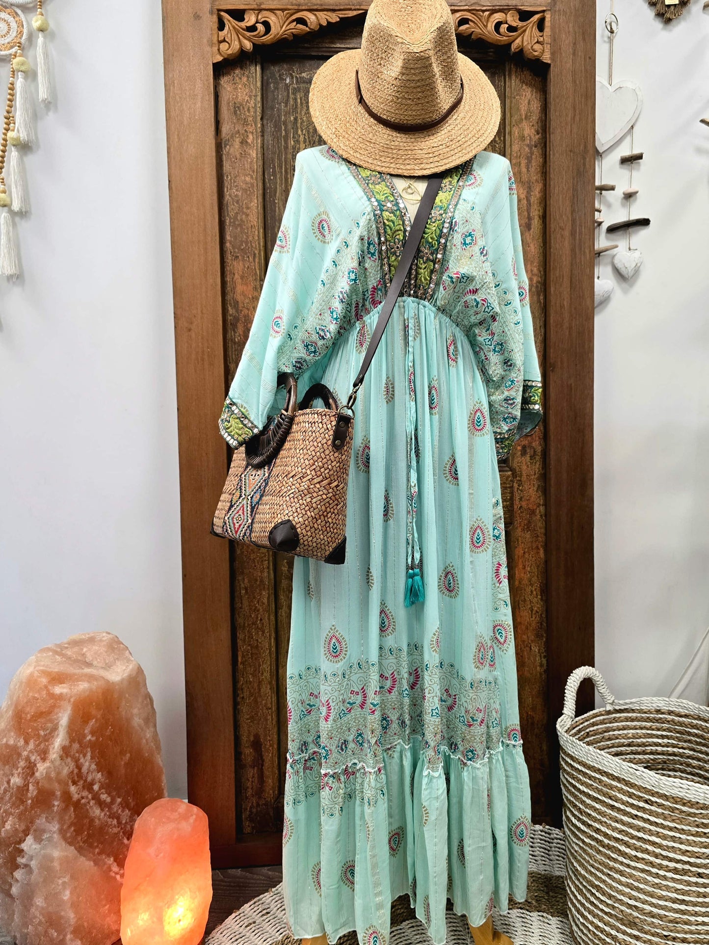 A bohemian-style light aqua maxi dress with colorful paisley and floral patterns, styled on a mannequin with a straw hat, a woven bucket bag, and standing in front of a carved wooden door.