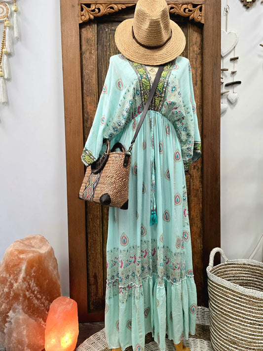 A bohemian-style light aqua maxi dress with colorful paisley and floral patterns, styled on a mannequin with a straw hat, a woven bucket bag, and standing in front of a carved wooden door.