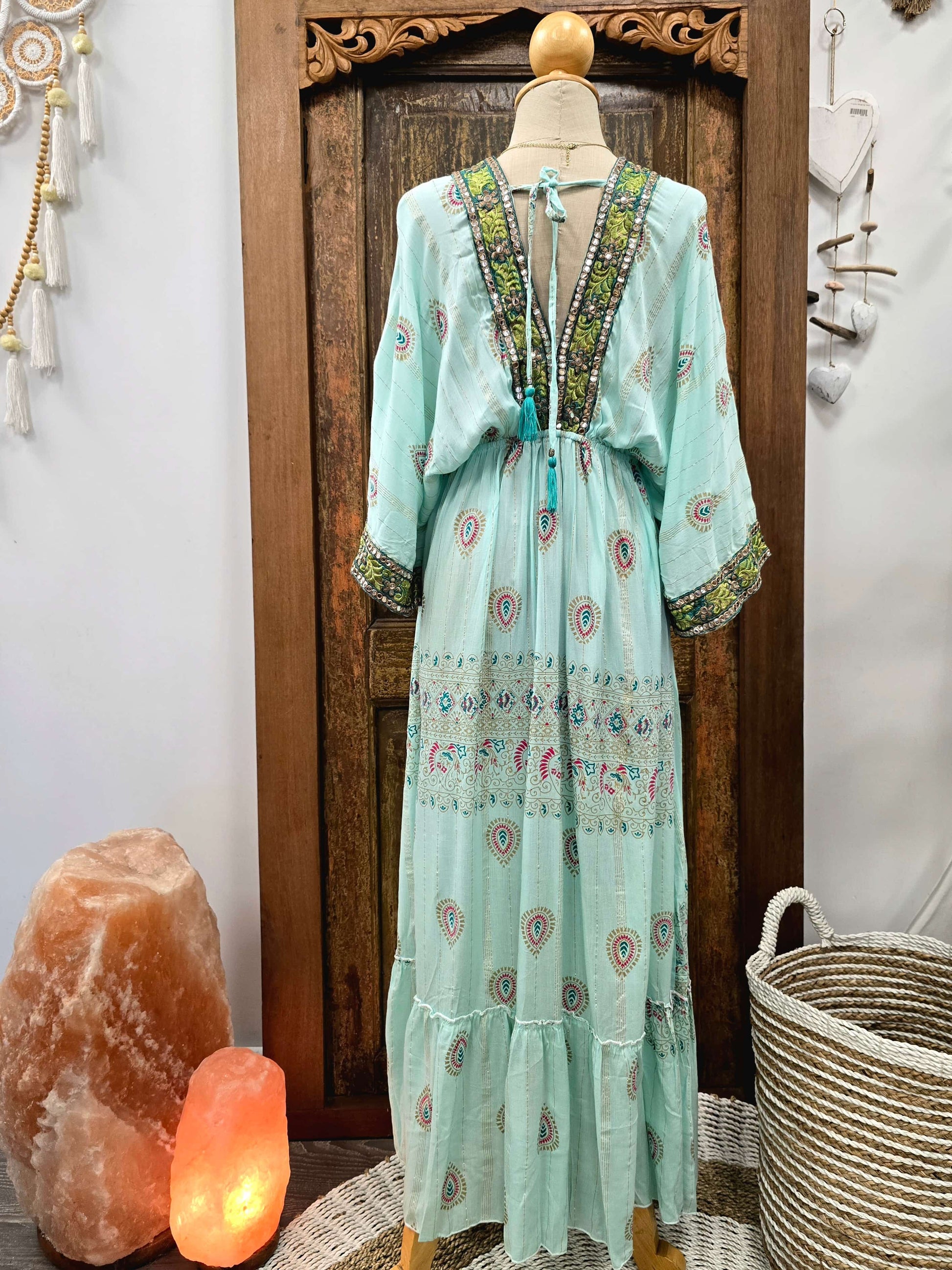 Back view of a light mint green bohemian maxi dress with colorful paisley and floral embroidery, displayed on a mannequin in front of a dark wooden door. The dress features a tie-neck design with tassel details and wide sleeves.