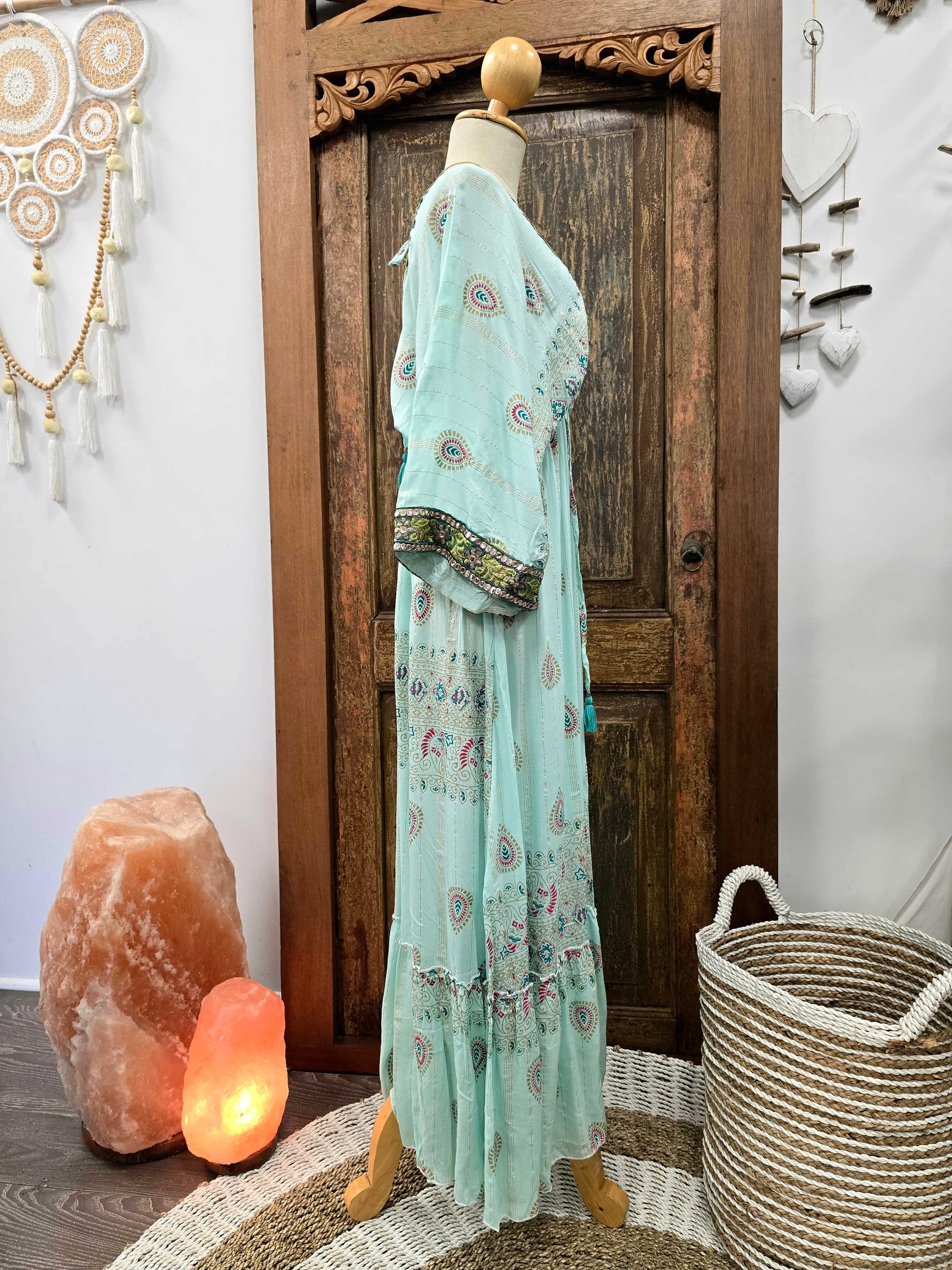 Side view of a light mint green bohemian maxi dress with colorful paisley prints and a patterned border on the sleeves, displayed on a wooden dress form in front of a rustic wooden door.