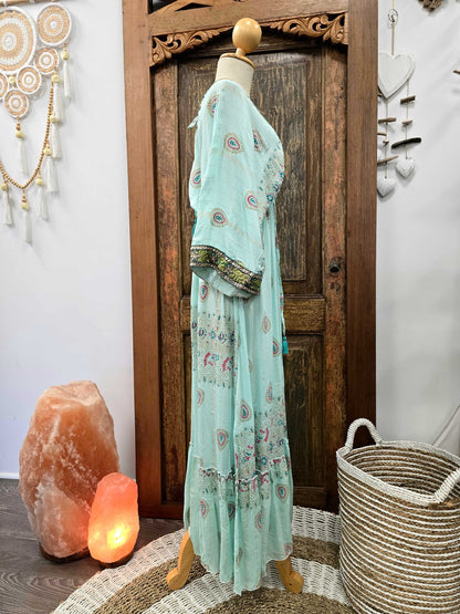 Side view of a light mint green bohemian maxi dress with colorful paisley prints and a patterned border on the sleeves, displayed on a wooden dress form in front of a rustic wooden door.