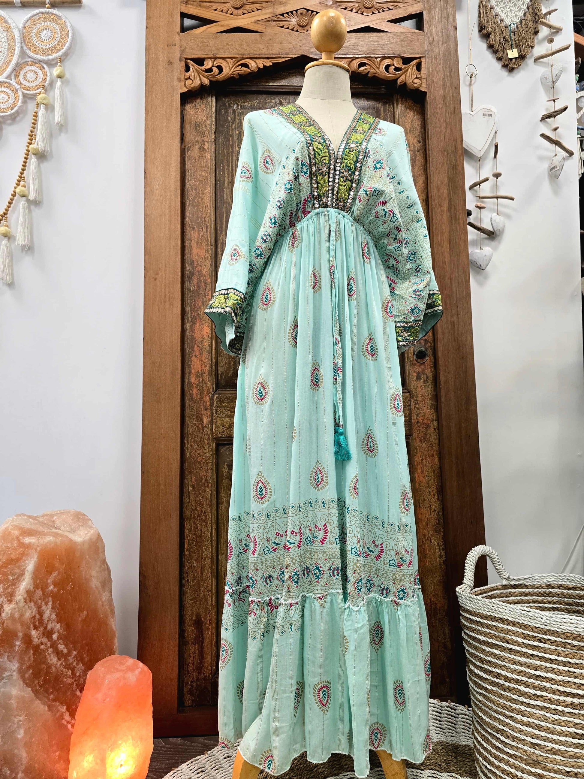 A long light aqua maxi dress with colorful paisley and floral embroidery, displayed on a mannequin in front of a carved wooden door. The dress features a V-neckline, long sleeves, and tiered ruffles.