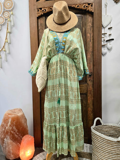 A bohemian-style long dress displayed on a mannequin, featuring a sage green and beige geometric pattern with turquoise blue beaded trim at the V-neck and cuffs. The dress is styled with a straw hat, a woven bag, and accessories against a rustic wooden door background.