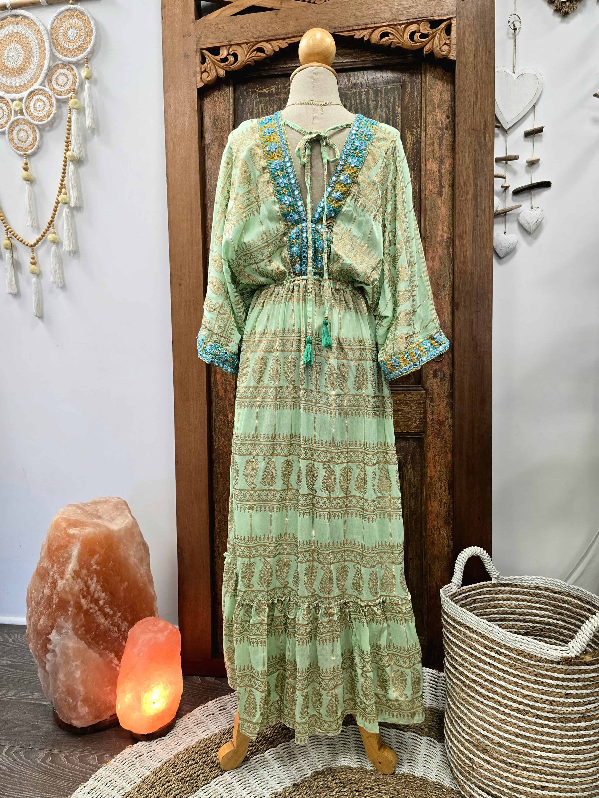 A light sage green bohemian maxi dress with blue and gold patterns, displayed on a mannequin in front of a rustic wooden door, flanked by a salt lamp, a woven basket, and wall decorations.