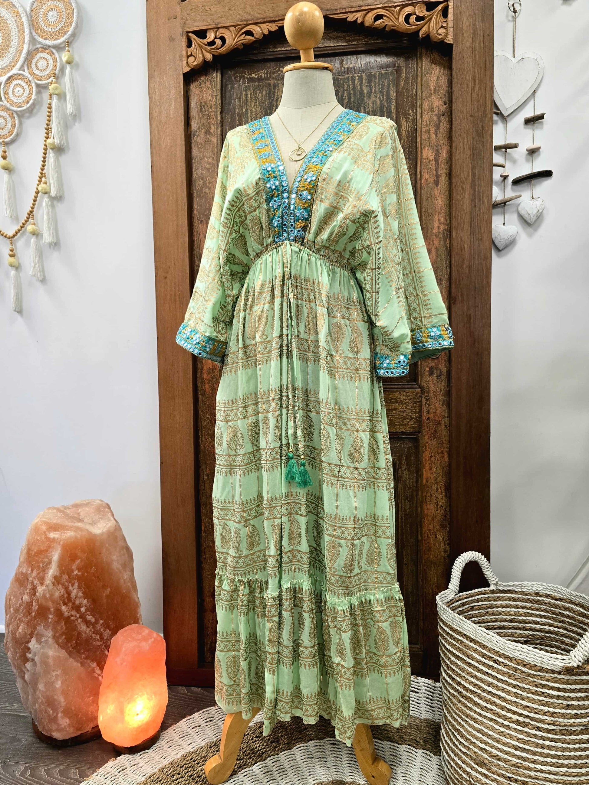 A light green bohemian maxi dress with blue and gold printed patterns, displayed on a mannequin in front of a wooden door, flanked by a salt lamp and a woven basket.