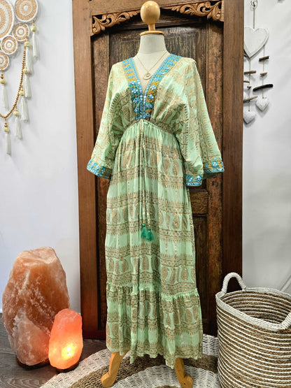 A light green bohemian maxi dress with blue and gold printed patterns, displayed on a mannequin in front of a wooden door, flanked by a salt lamp and a woven basket.