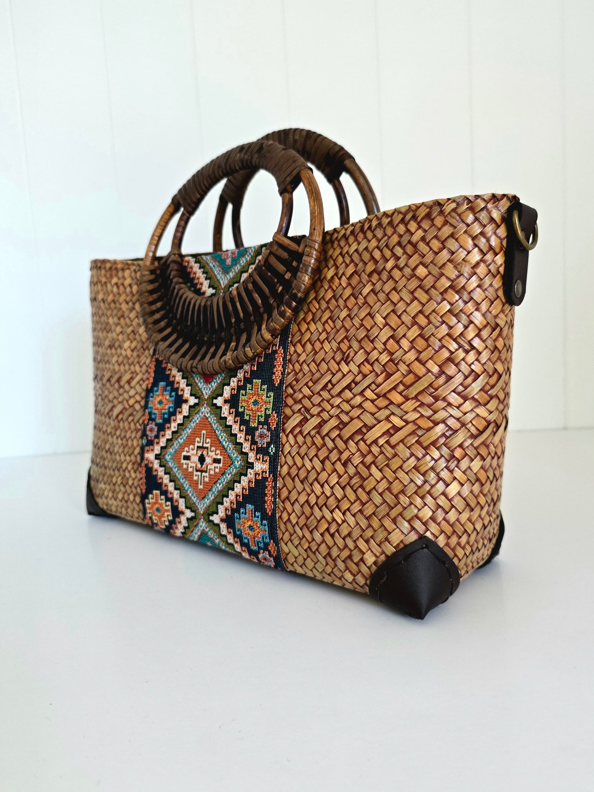 A woven straw tote bag with dark brown circular rattan handles, black faux leather corner protectors and strap, and a colorful geometric patterned panel on the front.