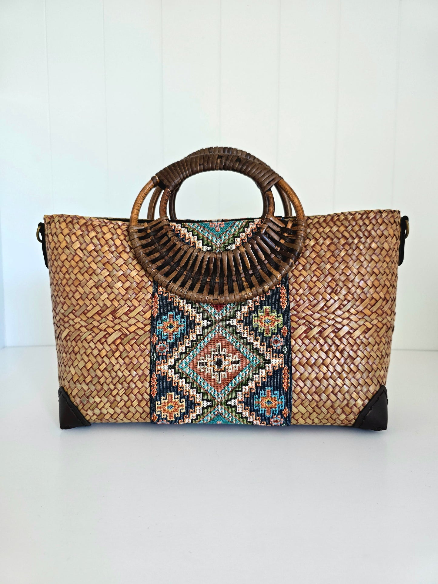A woven straw tote bag with a circular bamboo handle, featuring a colorful geometric pattern panel in the center and black protective corner guards, set against a white paneled background.