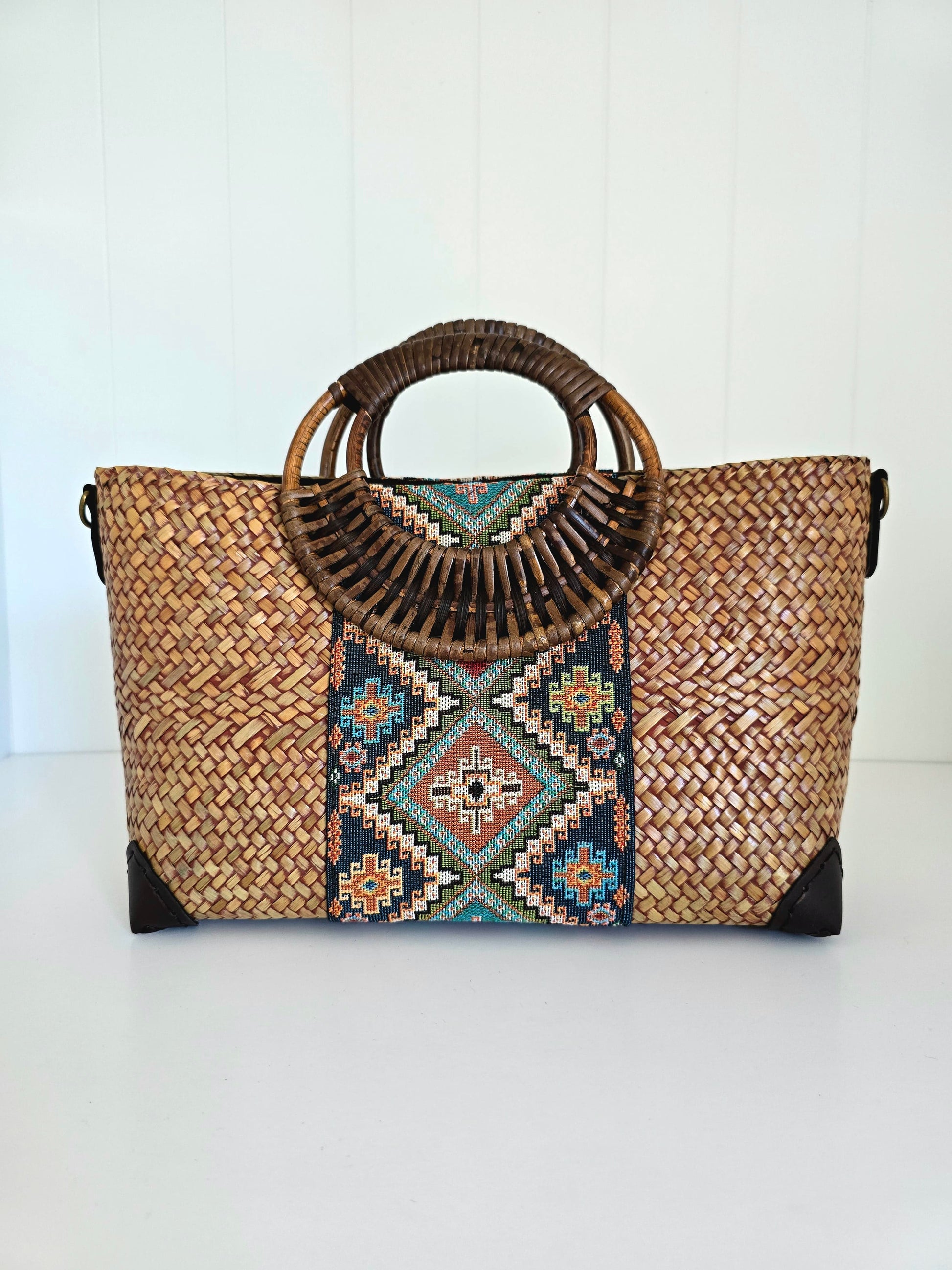 A woven straw tote bag with a circular bamboo handle, featuring a colorful geometric pattern panel in the center and black protective corner guards, set against a white paneled background.