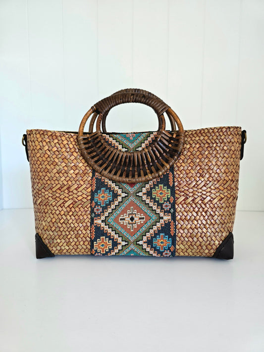 A woven straw tote bag with a circular bamboo handle, featuring a colorful geometric pattern panel in the center and black protective corner guards, set against a white paneled background.