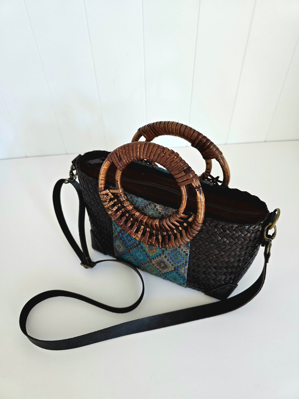 A woven black and brown handbag with two large circular rattan handles, a blue patterned fabric panel, and a long black shoulder strap, photographed on a white surface against a white paneled background.