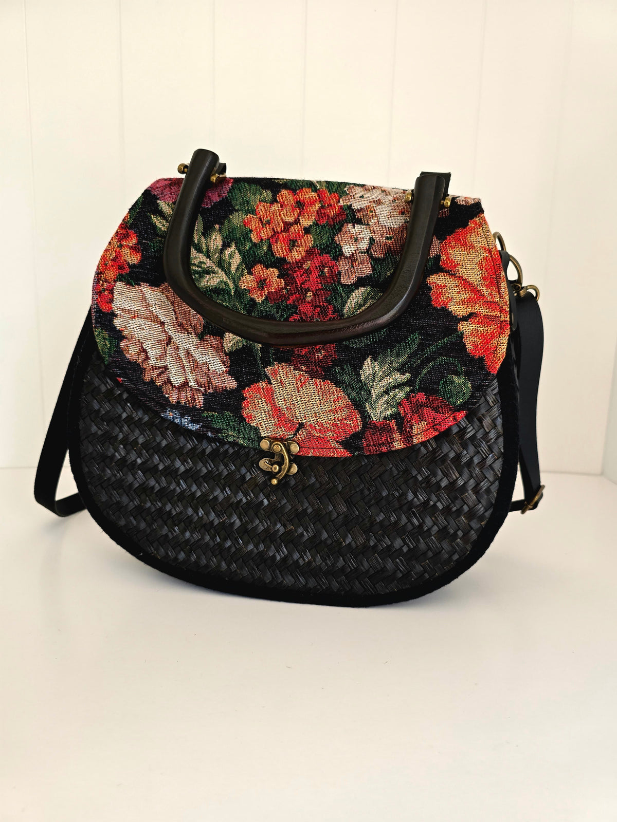 A black woven handbag with a colorful floral tapestry top panel and wooden handle, shown on a white background.