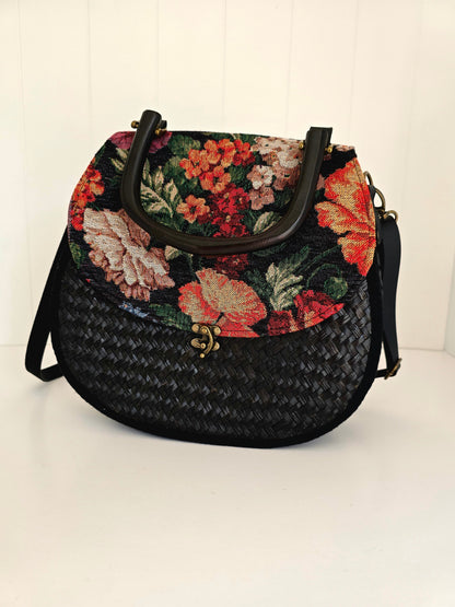 A black woven handbag with a colorful floral tapestry top panel and wooden handle, shown on a white background.
