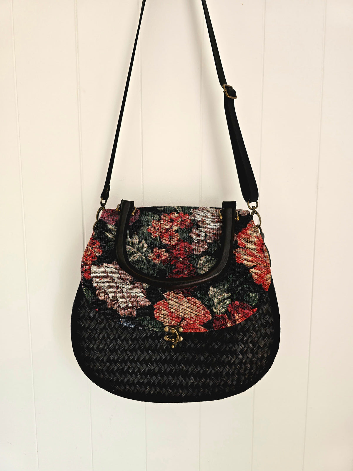 A black woven shoulder bag with a floral patterned tapestry panel and black strap, hanging against a white paneled wall.