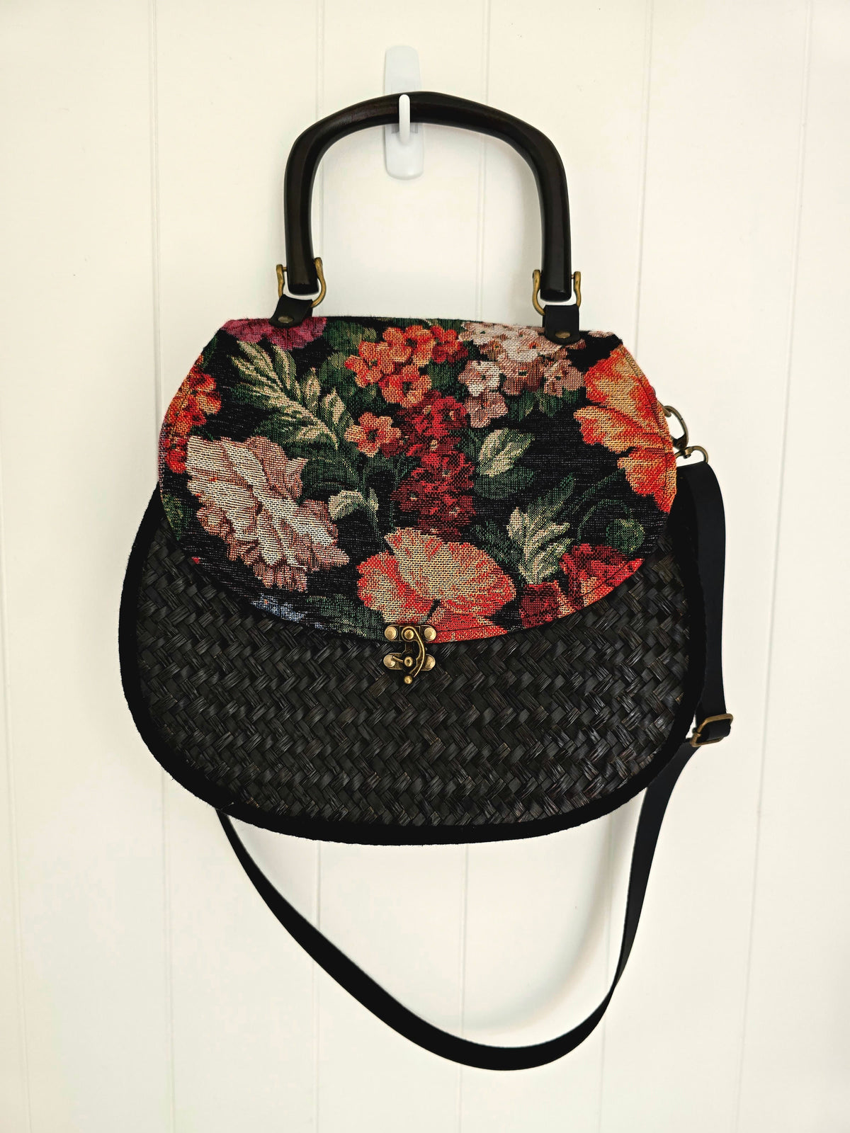 A vintage-style handbag with a black woven body, black faux leather shoulder strap and wooden top handle, and a floral tapestry upper panel in red, pink, and green, hanging against a white paneled wall.
