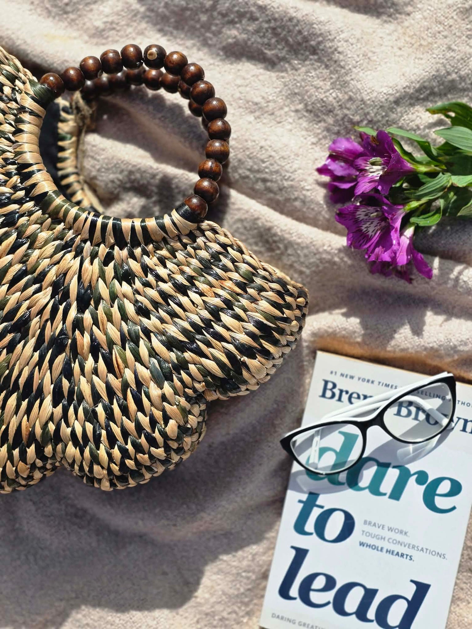 A woven straw handbag with a dark brown wooden bead handle, styled on a beige textured blanket with a book, eyeglasses, and purple flowers.