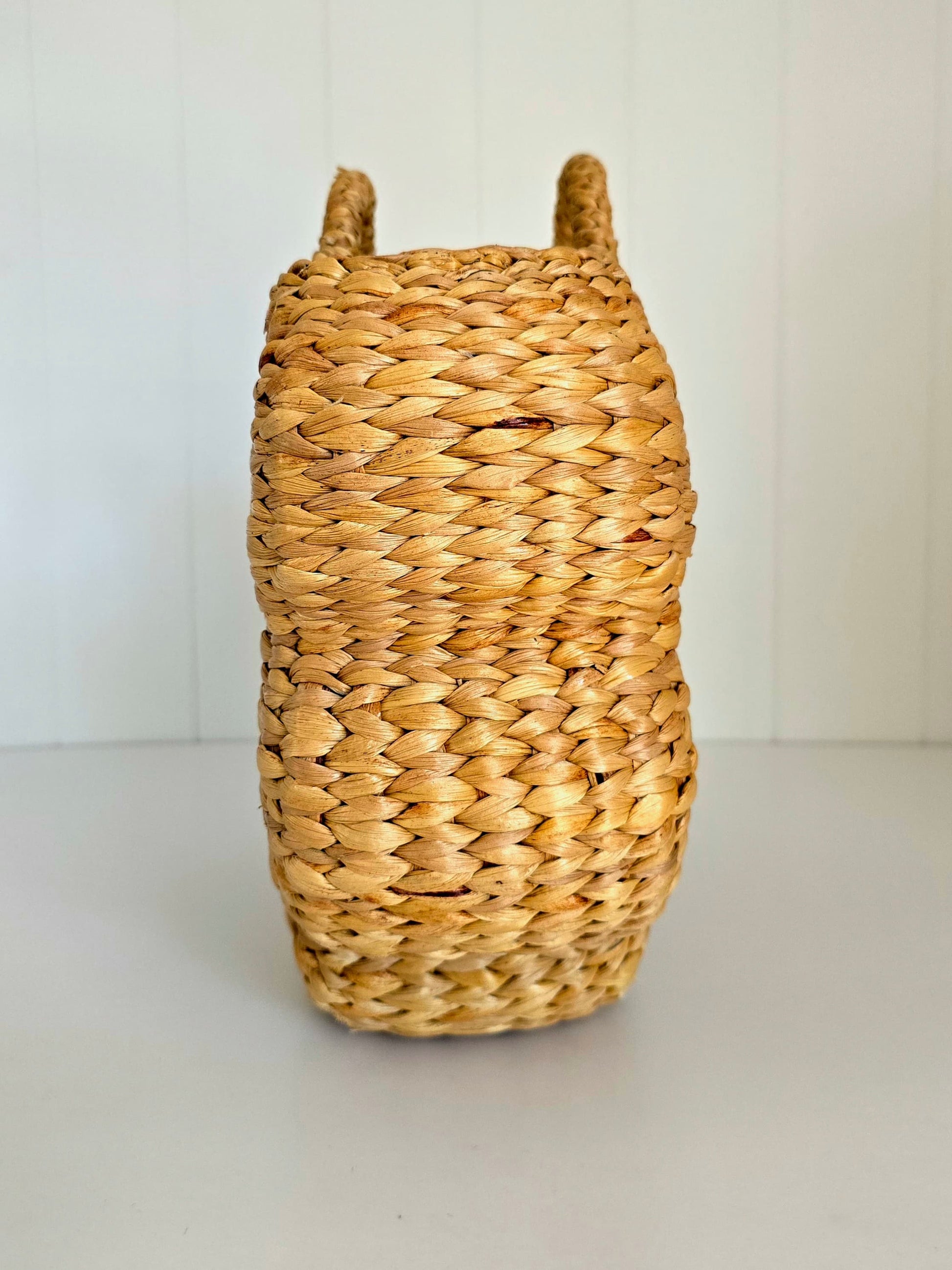 A natural woven straw basket with two upright handles, photographed against a clean white paneled background.