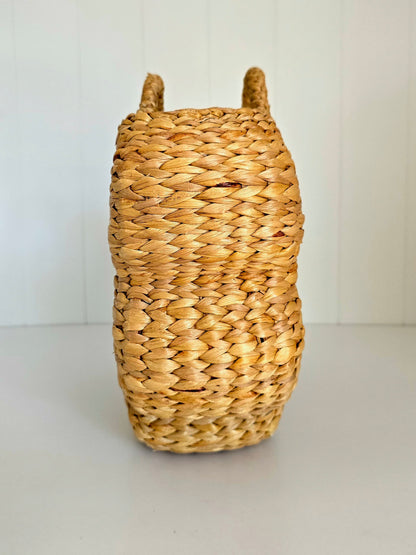 A natural woven straw basket with two upright handles, photographed against a clean white paneled background.