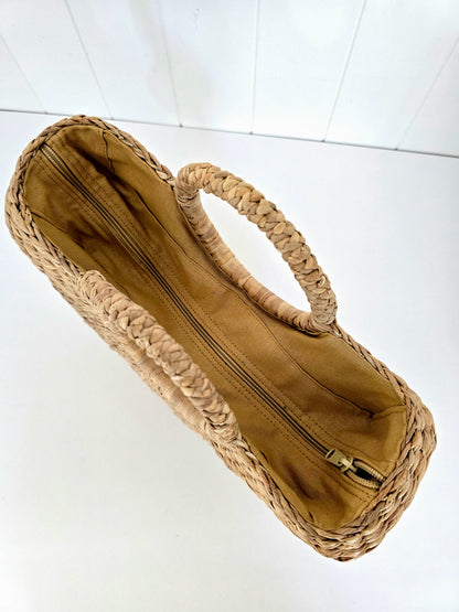 A woven straw handbag with a tan fabric lining and a single integrated top handle, shown from an overhead angle on a white background.