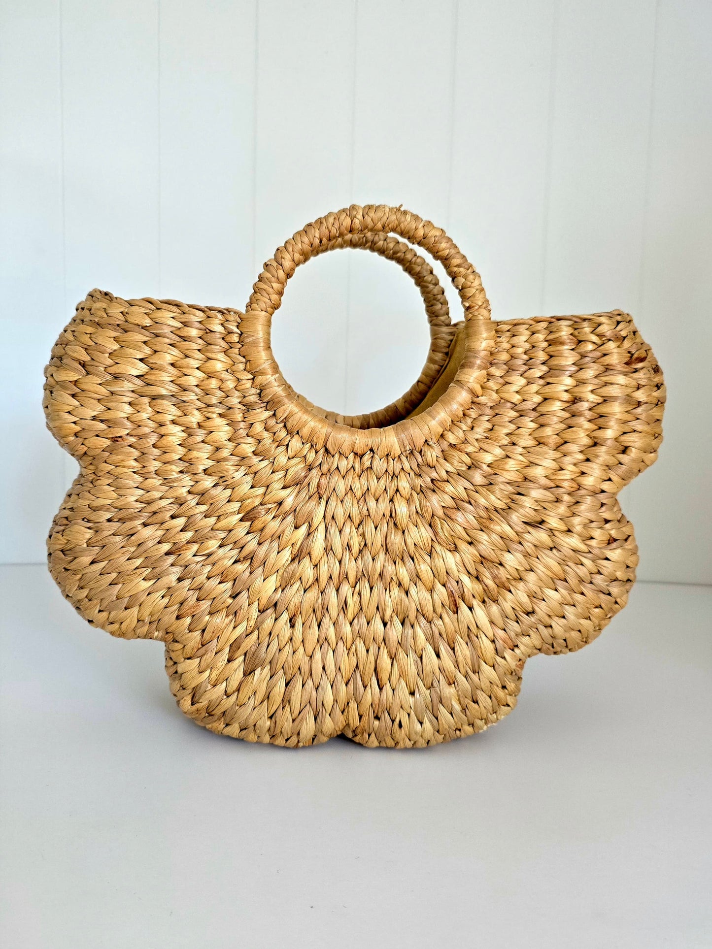A natural hand woven straw tote bag with a scalloped edge and a single integrated circular handle, shown against a white paneled background.