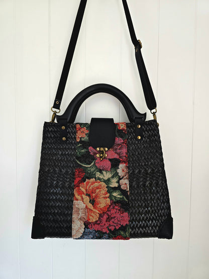 A black woven handbag with a floral tapestry panel on the front, black faucx leather shoulder strap and wooden handle, and gold-tone hardware, hanging against a white paneled wall.