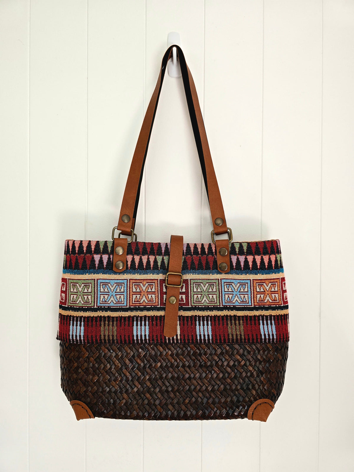 A woven tote bag with a brown leather shoulder strap and trim, featuring a geometric patterned band and a dark brown woven body, hanging against a white paneled wall.