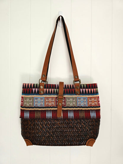 A woven tote bag with a brown leather shoulder strap and trim, featuring a geometric patterned band and a dark brown woven body, hanging against a white paneled wall.