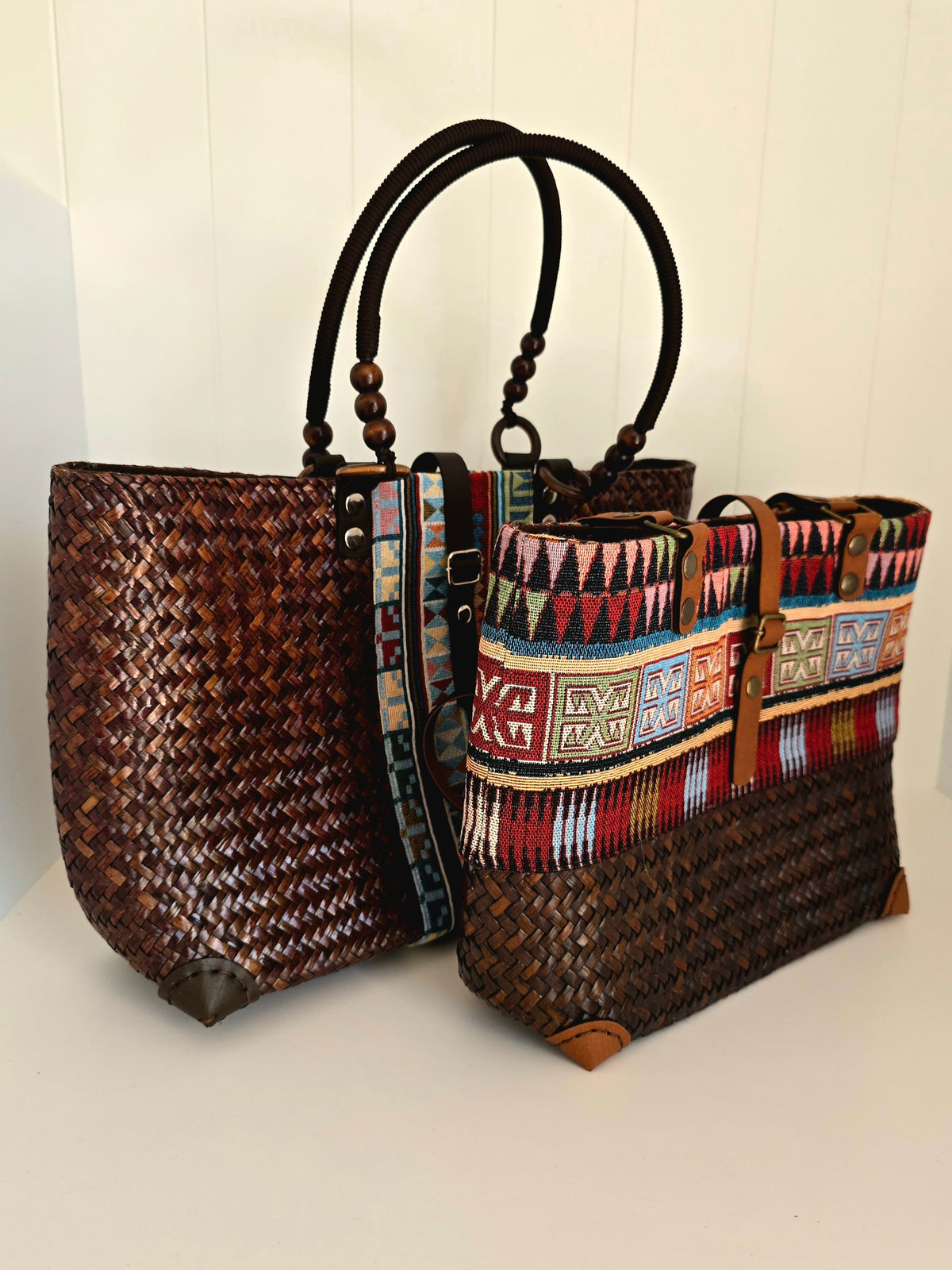 Two woven tote bags displayed together on a white surface against a white paneled wall. The left bag is made of dark brown woven material with black handles and dark trim.