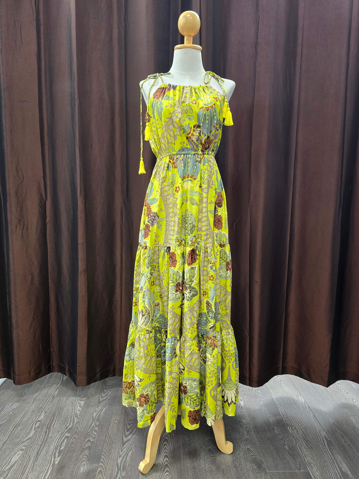 A sleeveless, tiered maxi dress with a vibrant yellow and green botanical print, displayed on a wooden mannequin in front of a dark brown curtain backdrop.