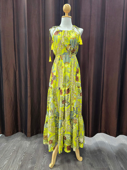 A sleeveless, tiered maxi dress with a vibrant yellow and green botanical print, displayed on a wooden mannequin in front of a dark brown curtain backdrop.