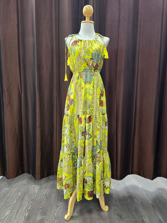 A sleeveless, tiered maxi dress with a vibrant yellow and green botanical print, displayed on a wooden mannequin in front of a dark brown curtain backdrop.