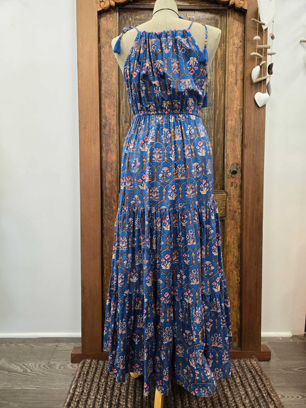 A sleeveless tiered maxi dress with a blue base and a red, white, and yellow floral/anchor pattern, displayed on a mannequin in front of a rustic wooden door.