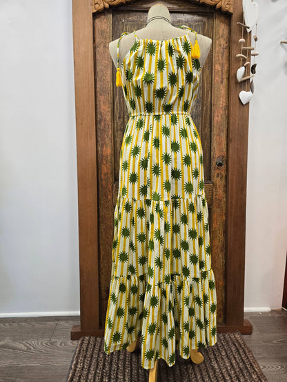 A sleeveless tiered maxi dress with a yellow and white vertical stripe pattern and green palm tree print, displayed on a mannequin in front of a rustic wooden door.